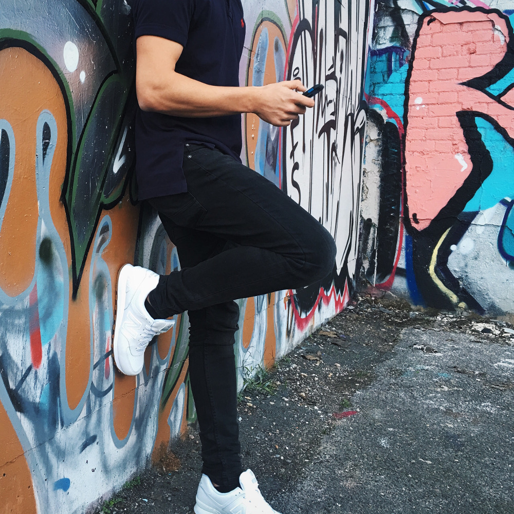 homme adossé contre un mur taggé portant un polo un jean noir et des baskets blanches